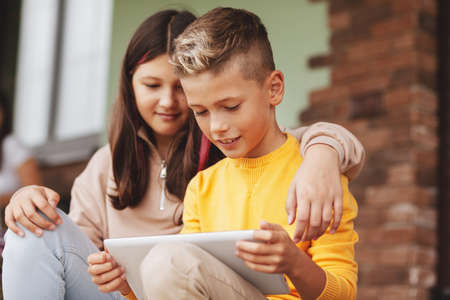 Brother and sister sit on the steps after school and play on a digital tablet. Modern children,...