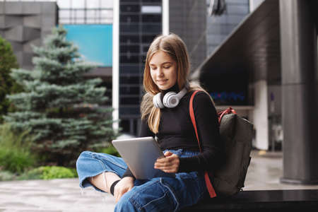 Beautiful fashionable schoolgirl studying online with tablet and headphones sitting on the steps near the building, side viewの写真素材