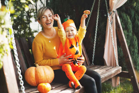 A young mother and her little daughter in a Halloween costume are sitting on a swing in the garden, next to orange pumpkins. The concept of the holiday, Halloweenの写真素材