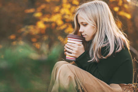 A beautiful teenage girl with blond hair sits thoughtfully in an autumn park, holds a flask with tea...