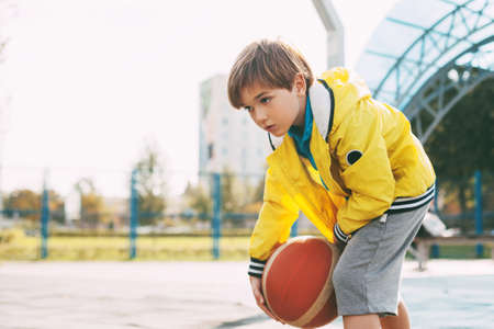 A cute boy in a yellow sports jacket throws a basketball. A child plays basketball. Sports, training, lifestyleの写真素材