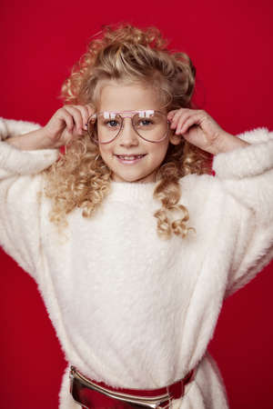 A portrait of a little curly blonde in a white fluffy sweater and a red cap with a scarf stands and smiles on a red background. Festive postcard. Merry Christmas and Happy New Year.の写真素材