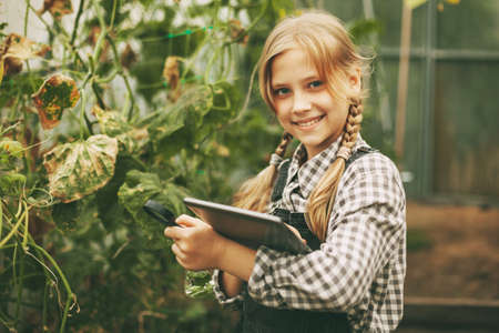 A beautiful little teenage girl with a tablet in her hands examines a sample of a plant through a magnifying glass. Curious children. Organic pure productsの写真素材