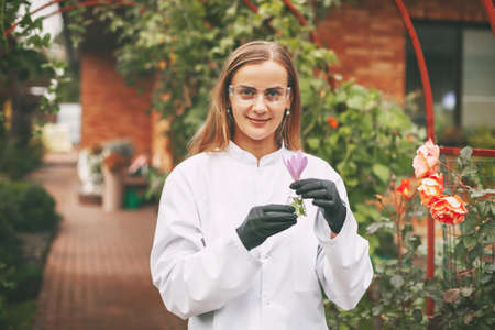 A woman scientist in a white coat and glasses examines a sample of a plant through a magnifying...
