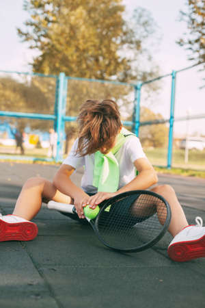 Cute teen sitting on the tennis court and resting after a hard workout. Sport, sportsman, lifestyle, leisureの写真素材
