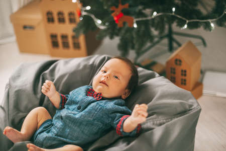 A small child lies on a pouf next to a Christmas tree and smiles. The kid is happy with the holiday....