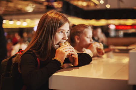 Beautiful schoolgirl eating hamburger sitting in a cafe indoors. Happy child eating junk food in a restaurant. Side viewの写真素材