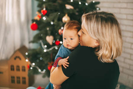 A mother holds her little son in her arms and kisses him . Baby with mom in a festively decorated...