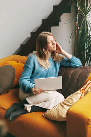 Pensive beautiful girl sitting on the couch with a laptop. Young woman working at home using laptopの写真素材