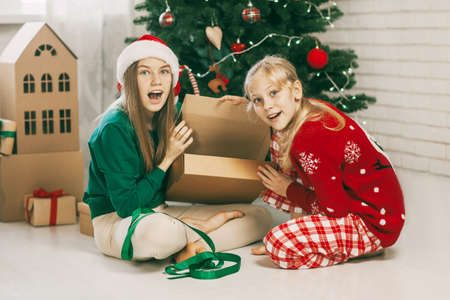 Two happy and cheerful sisters in fashionable New Year's outfits open gifts at home under the Christmas tree. Christmas mood. Concept for New Years holiday at homeの写真素材