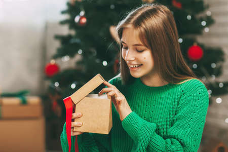 Beautiful teenage girl opens a gift made of kraft paper tied with a red ribbon, new year mood....