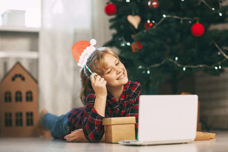 A happy little girl opens a gift under the Christmas tree at home and communicates with her grandparents via a laptop via video link. Holiday concept for Christmas and New Year at homeの写真素材