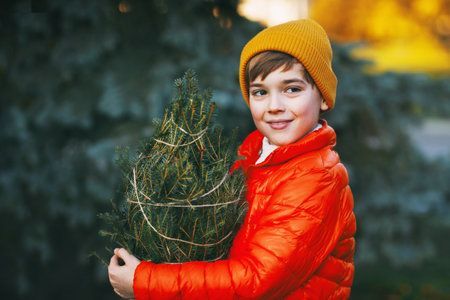 A boy in a bright orange jacket and yellow hat holds a purchased Christmas tree in his hands, smiles and looks into the distance. Shopping for the holiday. Preparing for Christmas, New Yearの写真素材