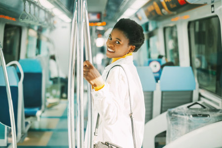 A smiling young African woman holds on to a handrail while standing on a subway train during her daily commute to workの写真素材