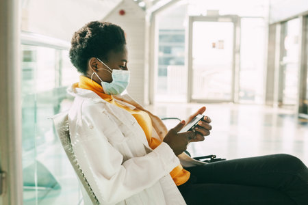 A young African woman in a protective mask sits in a chair in the waiting room and reads emails on her phone. Social distance, pandemic.の写真素材