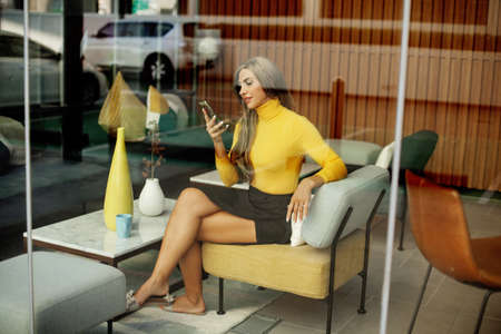 A young woman drinks coffee and uses her smartphone while sitting in a city cafe. Modern business, business onlineの写真素材