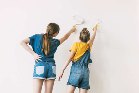 Two sisters are painting the walls of the house white, in denim overalls and a bright yellow...