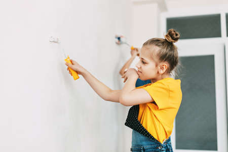 A girl in a denim overalls and a yellow T-shirt helps to paint the walls in an apartment whiteの写真素材