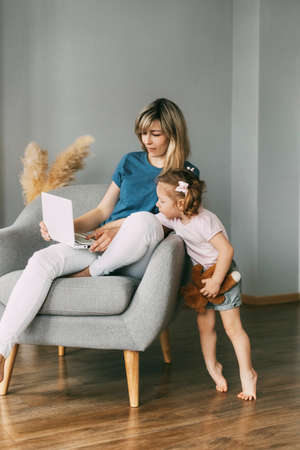 A young mother is working on a laptop sitting in a chair, and her little daughter is watching the work. Freelance, business online, Work onlineの写真素材
