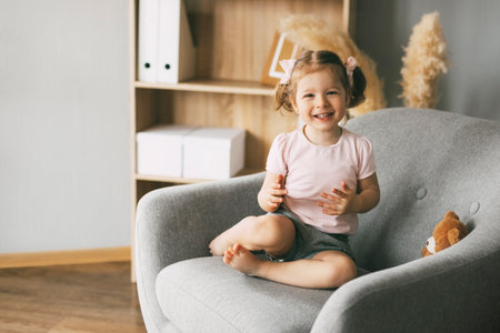 A little charming girl sits in a chair with her toy bear and smilesの写真素材