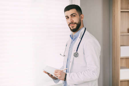 Modern young male doctor of oriental appearance stands in his office
