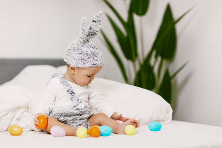 Baby sits on a bed in a bunny costume and plays with colorful easter eggs, laughs and has fun