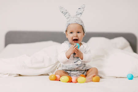 Baby sits on a bed in a bunny costume and plays with colorful easter eggs, laughs and has funの写真素材