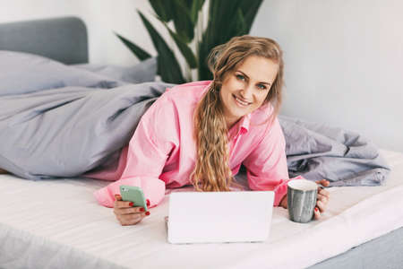 A beautiful girl is lying on the bed in a pink shirt, holding a cup of coffee in her hand and using a laptop to view online content, online training, work onlineの写真素材