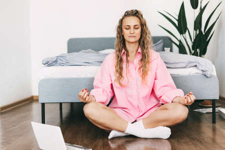 A young woman meditates while sitting next to a bed in . The girl does yoga exercises at home to relieve stress, taking a break from work.の写真素材