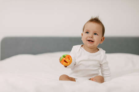 The kid sits in a snow-white bed, laughs and plays with a rattle.