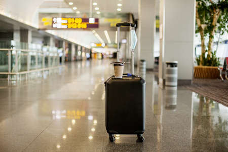 In an empty airport, there is a suitcase, a glass of coffee on it, and a medical protective mask.の写真素材