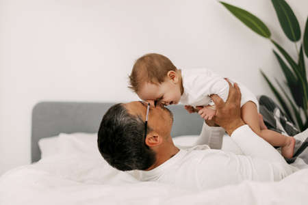Happy father playing with adorable baby in the bedroom in a snow-white bed