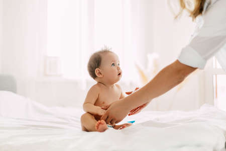 The cute baby is sitting on the bed, holding her mothers hands and looking at her. Care, baby, motherhood, childhoodの写真素材