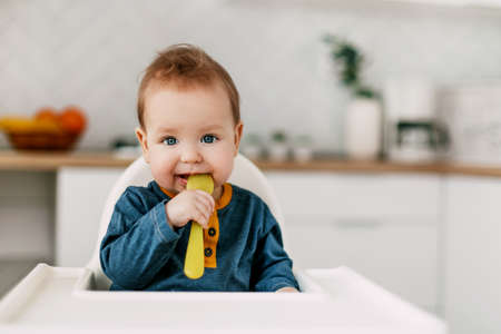 The baby is sitting in a high chair with a yellow spoon in the kitchenの写真素材
