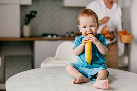 A little boy sits on the kitchen table with a banana in his hands and smiles