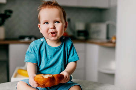 a charming baby sits on the kitchen table with a large bun in his handsの写真素材