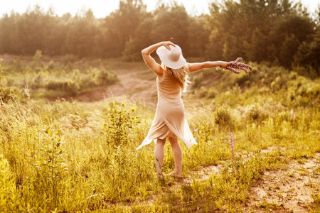 A young girl in a light dress and a hat with a bouquet in her hands is dancing on the field, looking at the sunset. Rear view, space for textの写真素材