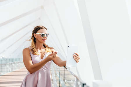 A girl in sunglasses and a summer dress communicates with friends using a tablet, making a video call while walkingの写真素材