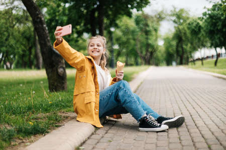 A blonde sitting on the road with a phone and ice cream makes a funny selfie. Child outdoors