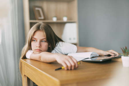 A teenage girl looks thoughtfully into the distance while doing her homework alone. The concept of...