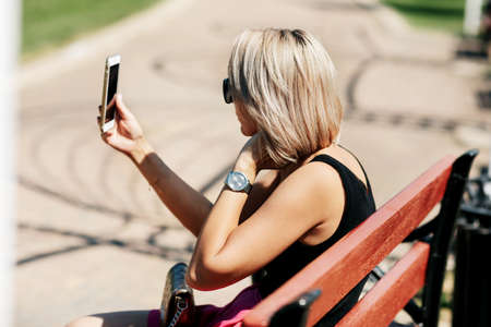 A young woman takes a selfie sitting on a park bench, adjusts her makeup and hairstyle, looking at her reflection on the screenの写真素材