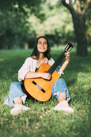 A laughing girl plays the guitar and sits on the grass in the park. Hobbies, music, musicianの写真素材