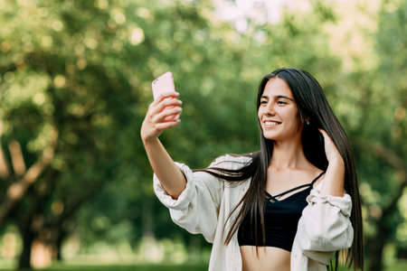 A young girl with long brown hair takes a selfie on a smartphone in the parkの写真素材