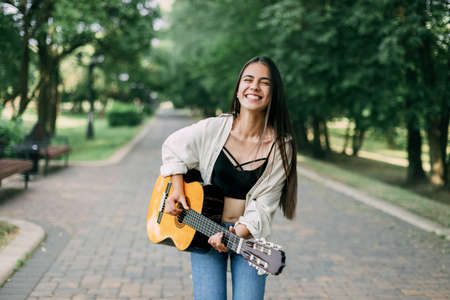 A cheerful musician girl enjoys playing the guitar in the park and laughs. Music, musician, guitar,...