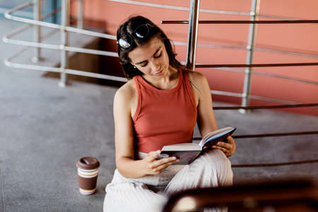 A student sits on the stairs on campus and reads a book, next to her is a glass of coffeeの写真素材