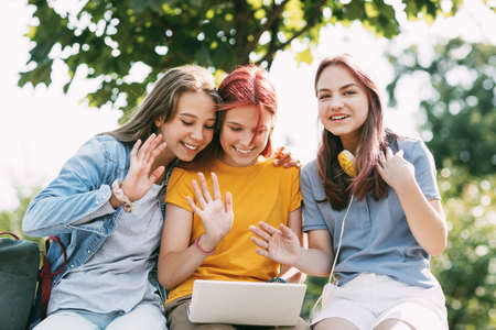 Three teenage girls communicate with their friends and classmates via video link using a laptop in...