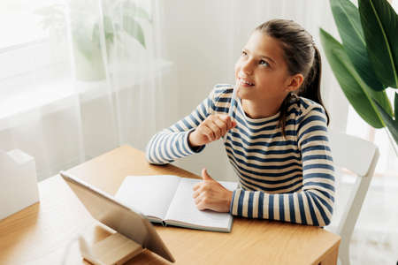 Happy pensive schoolgirl sits at the table at home, does homework on the tablet and looks out the...