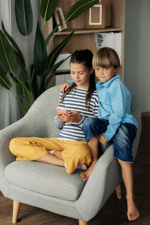 Sister and younger brother are sitting at home in an armchair and playing on the phone. Modern...