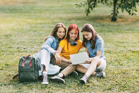 Teenage girls are sitting on a green lawn in a park with a backpack and a digital tablet and are preparing a joint project. Education and knowledge, lifestyle of teenagersの写真素材