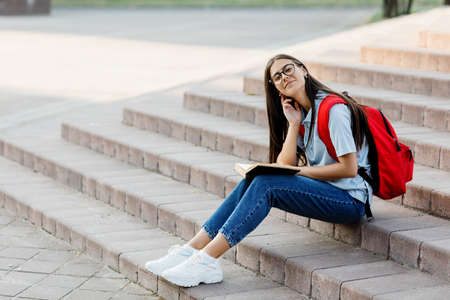 A young girl with glasses and a red backpack is sitting on the stairs with a book, spending time readingの写真素材